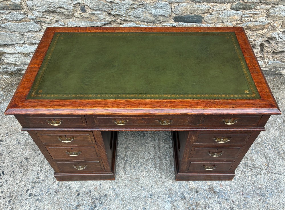 An Edwardian oak pedestal desk featuring a green leather writing surface, nine drawers with brass handles, and twin pedestals on plinth bases. View III