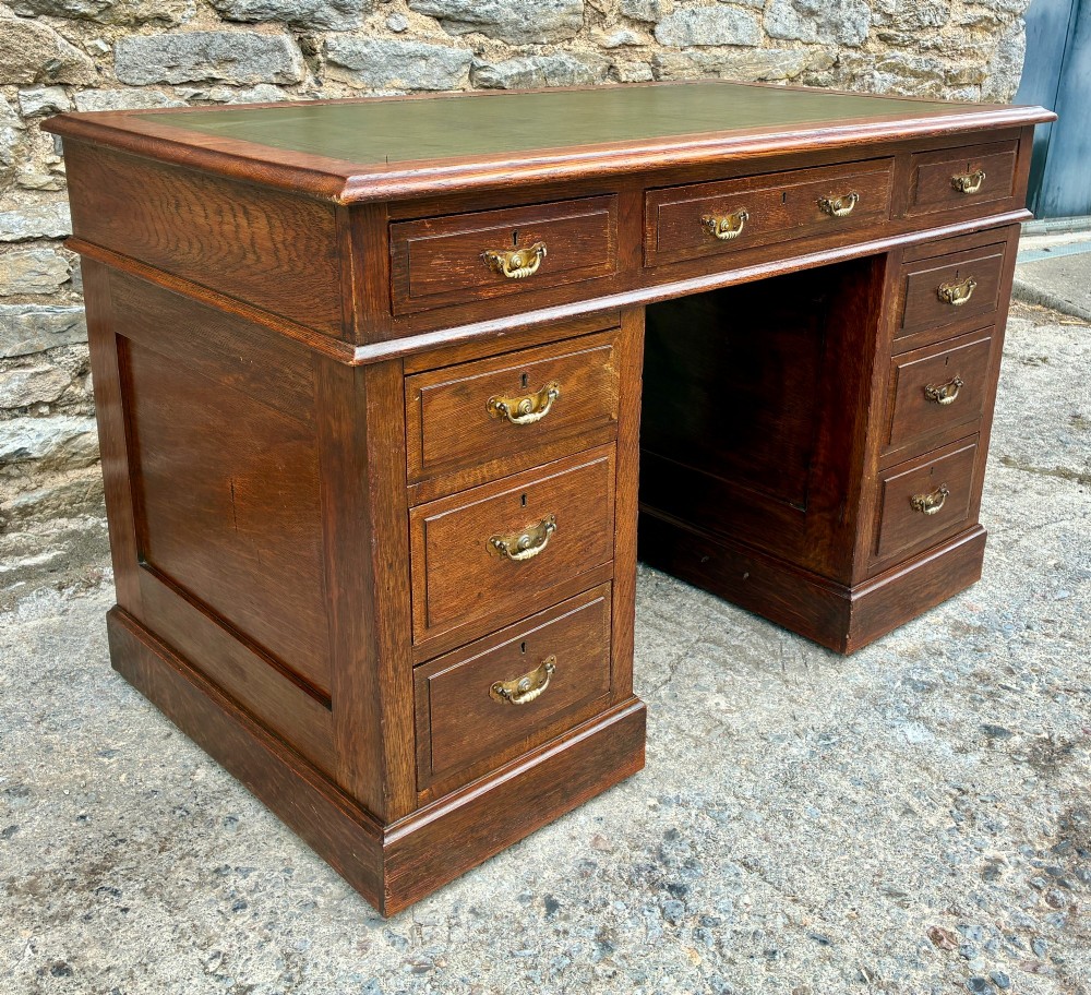 An Edwardian oak pedestal desk featuring a green leather writing surface, nine drawers with brass handles, and twin pedestals on plinth bases. View IV