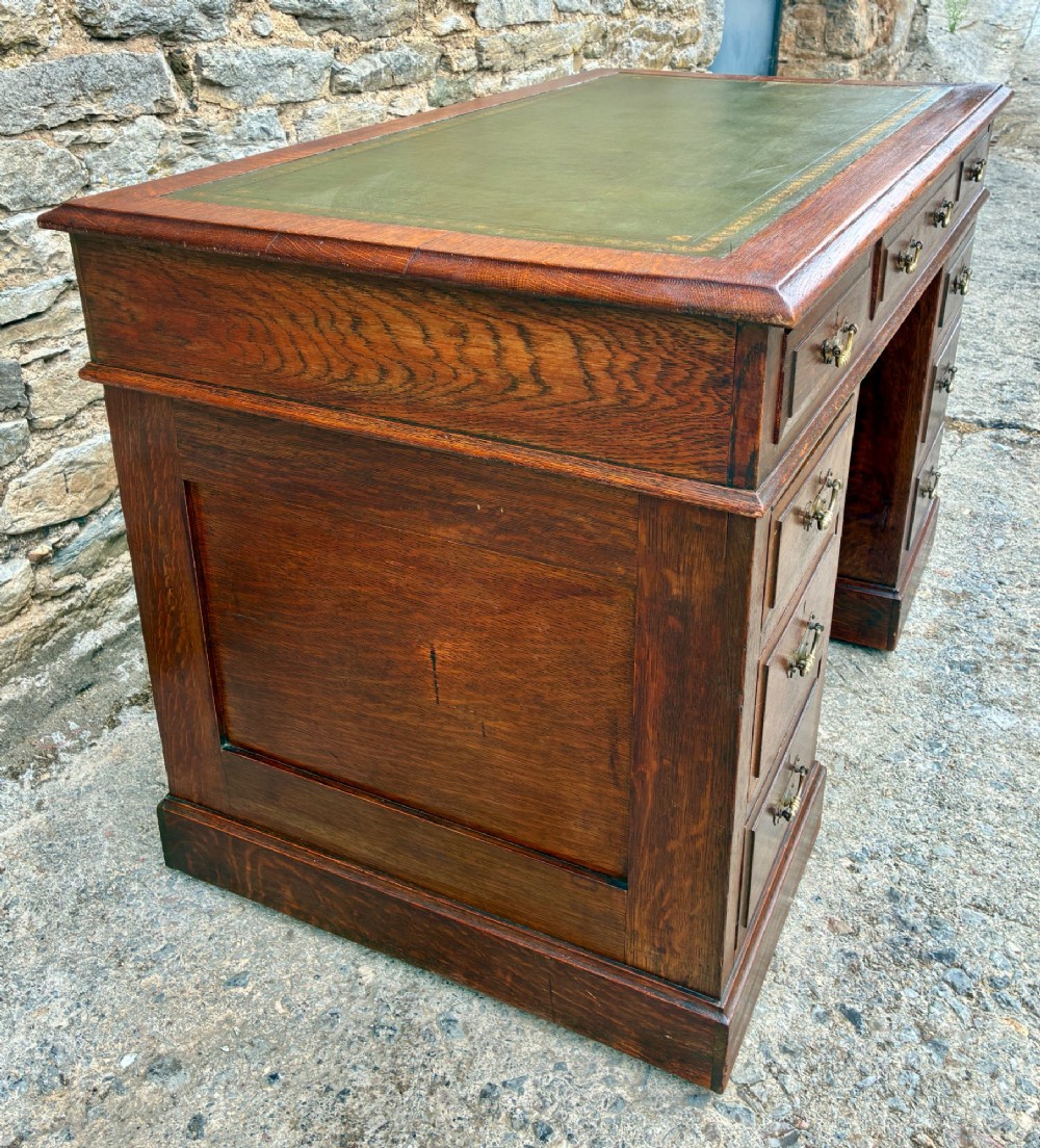 An Edwardian oak pedestal desk featuring a green leather writing surface, nine drawers with brass handles, and twin pedestals on plinth bases. View V