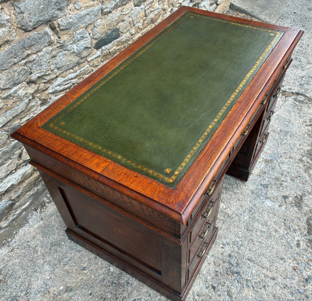 An Edwardian oak pedestal desk featuring a green leather writing surface, nine drawers with brass handles, and twin pedestals on plinth bases. View VI