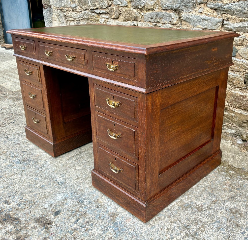 An Edwardian oak pedestal desk featuring a green leather writing surface, nine drawers with brass handles, and twin pedestals on plinth bases. View VII