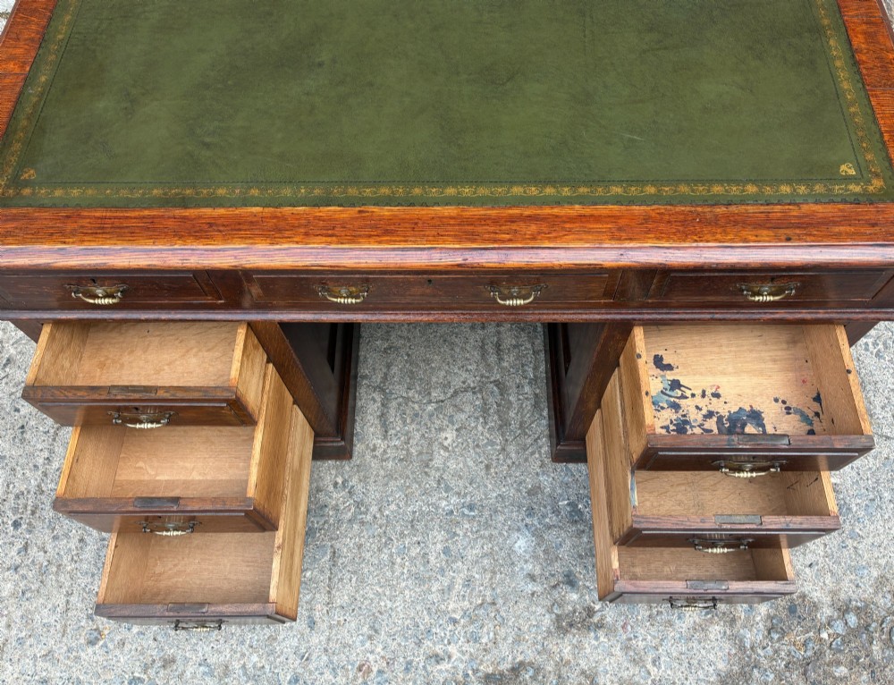 An Edwardian oak pedestal desk featuring a green leather writing surface, nine drawers with brass handles, and twin pedestals on plinth bases. View X