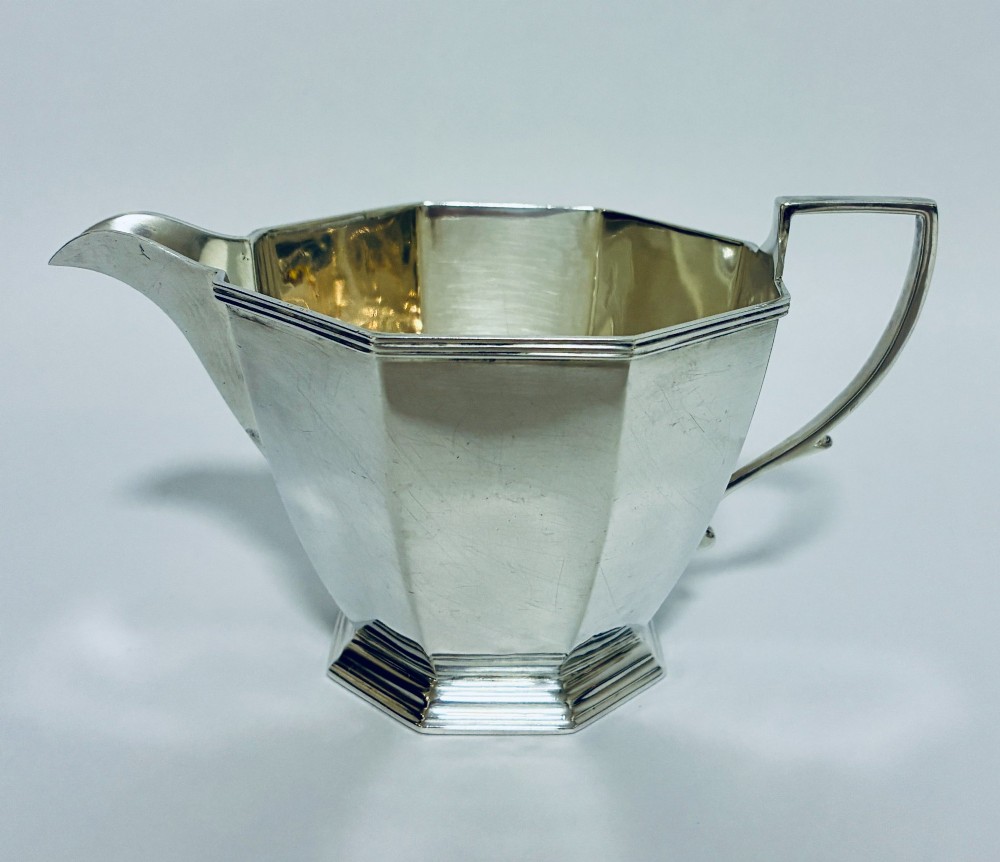 An octagonal sterling silver milk jug featuring an angular handle, a pronounced pouring spout, and visible stamped hallmarks on the front facet. View V