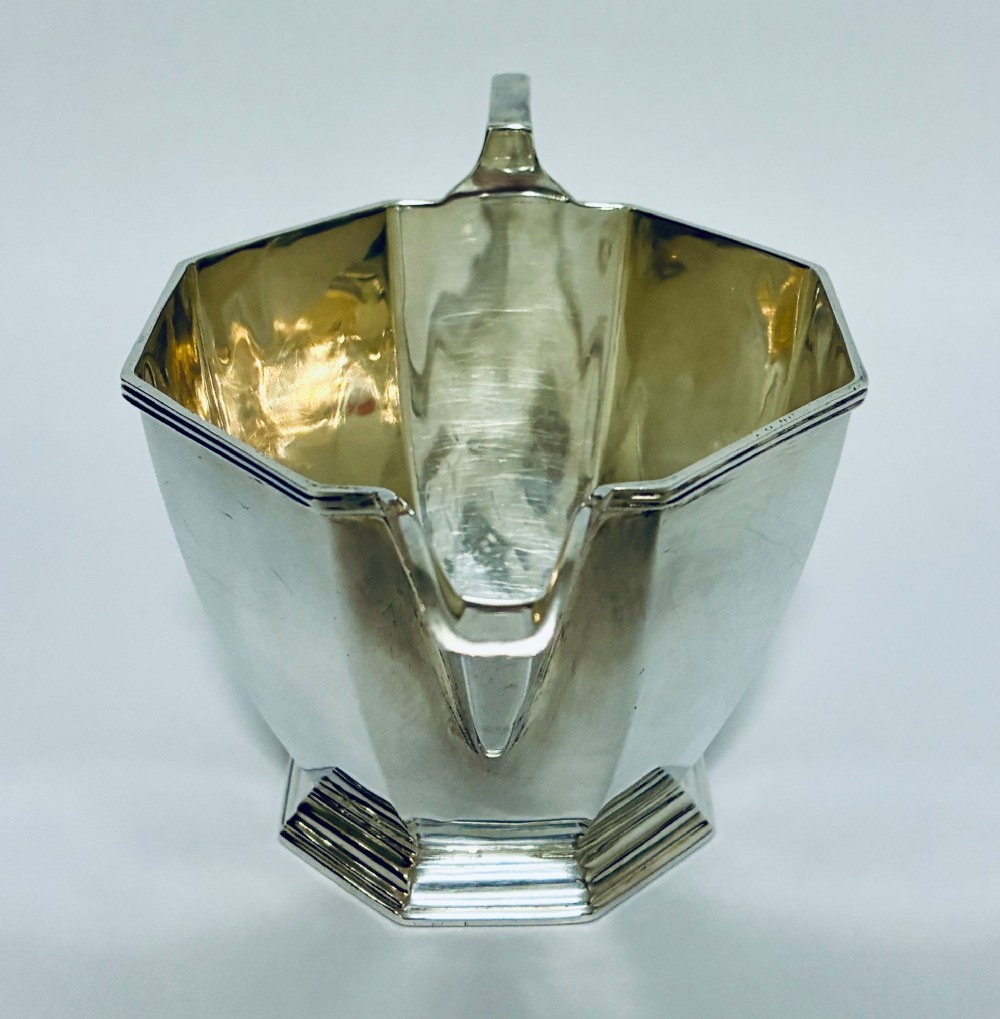 An octagonal sterling silver milk jug featuring an angular handle, a pronounced pouring spout, and visible stamped hallmarks on the front facet. View VII