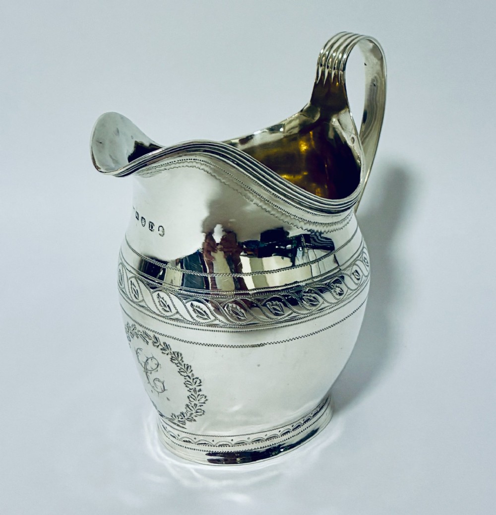 A Georgian sterling silver cream jug featuring a high reeded loop handle, engraved foliate bands around the body, and a gilded interior finish. View II