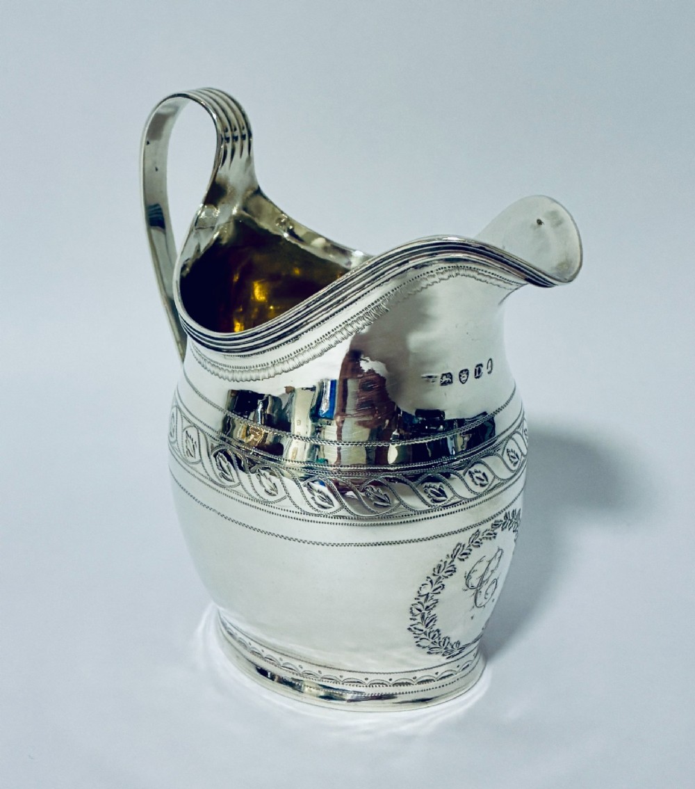 A Georgian sterling silver cream jug featuring a high reeded loop handle, engraved foliate bands around the body, and a gilded interior finish. View IV