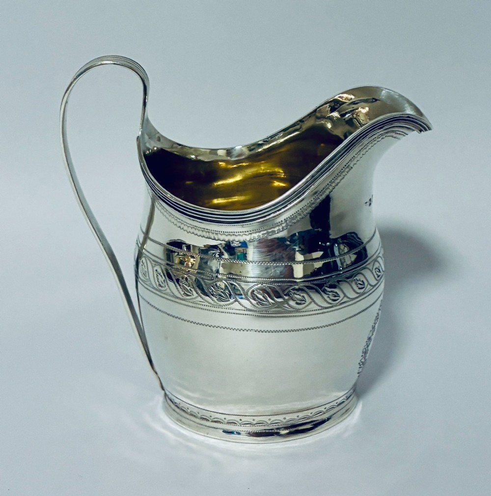 A Georgian sterling silver cream jug featuring a high reeded loop handle, engraved foliate bands around the body, and a gilded interior finish. View V