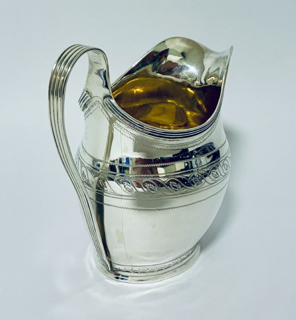A Georgian sterling silver cream jug featuring a high reeded loop handle, engraved foliate bands around the body, and a gilded interior finish. View VI