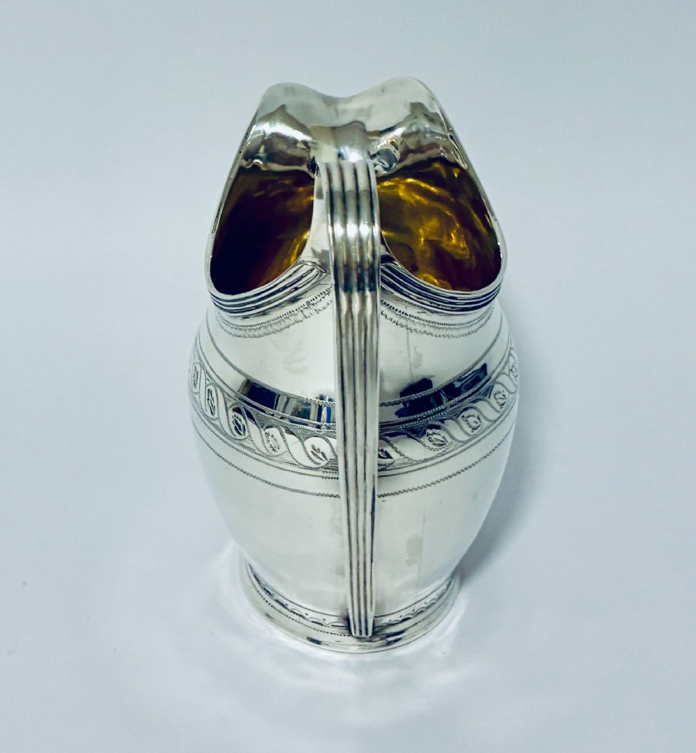 A Georgian sterling silver cream jug featuring a high reeded loop handle, engraved foliate bands around the body, and a gilded interior finish. View VII