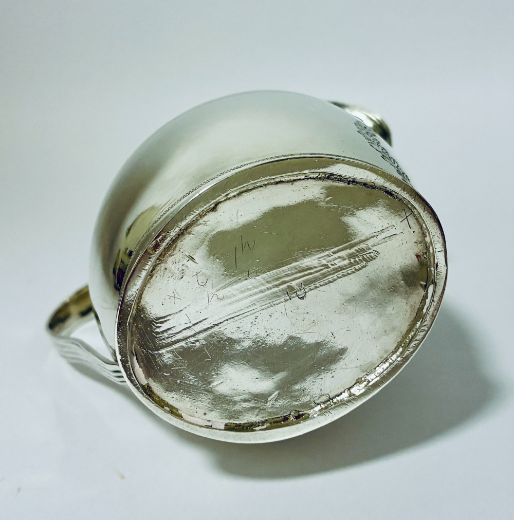 A Georgian sterling silver cream jug featuring a high reeded loop handle, engraved foliate bands around the body, and a gilded interior finish. View X