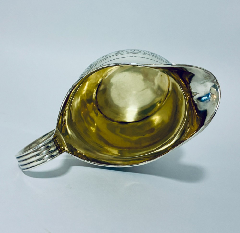 A Georgian sterling silver cream jug featuring a high reeded loop handle, engraved foliate bands around the body, and a gilded interior finish. View XI
