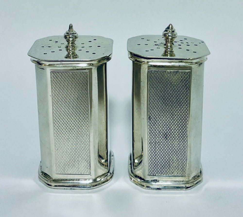 A pair of matching silver pepper pots with canted corners, engine-turned central panels, and perforated tops with decorative finials.