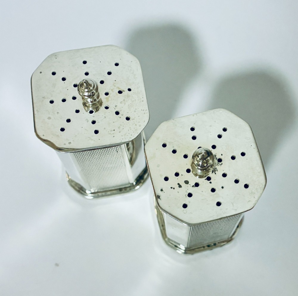 A pair of matching silver pepper pots with canted corners, engine-turned central panels, and perforated tops with decorative finials. View V