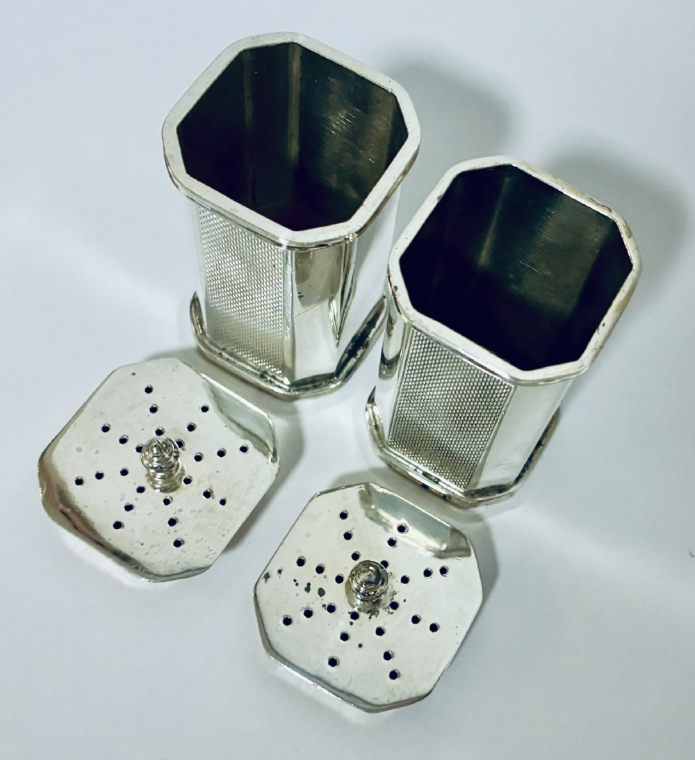 A pair of matching silver pepper pots with canted corners, engine-turned central panels, and perforated tops with decorative finials. View VI