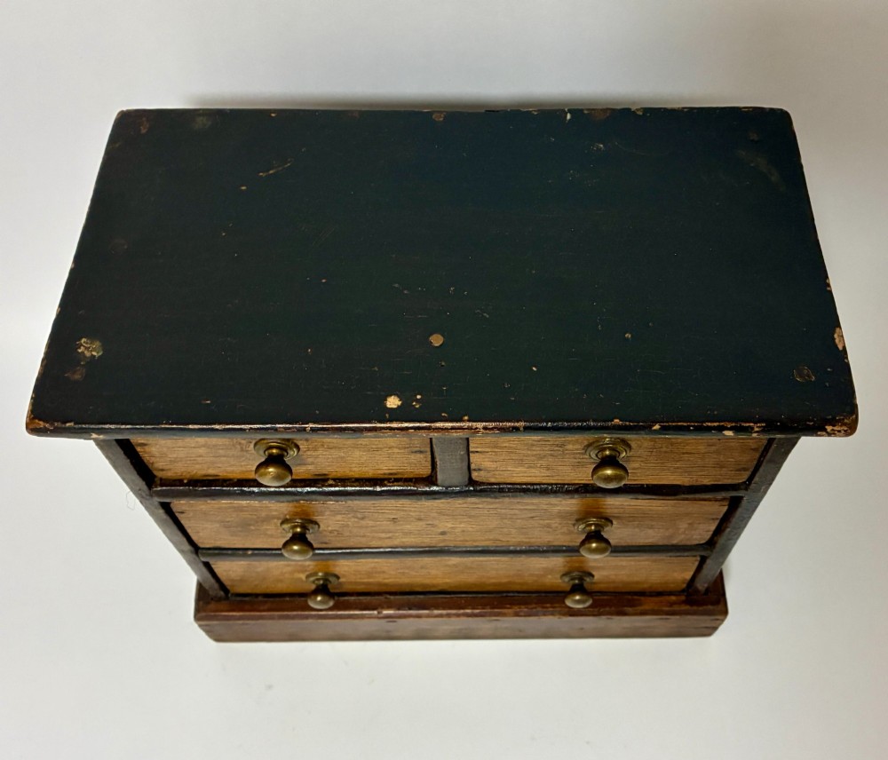 A small wooden miniature chest of drawers featuring two small upper drawers and two full-width lower drawers with turned brass knobs. View II