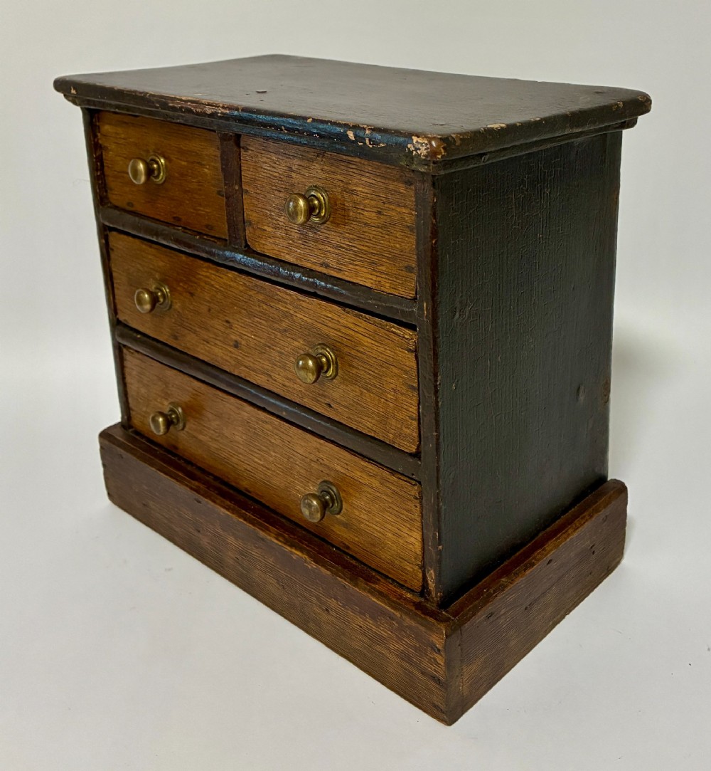 A small wooden miniature chest of drawers featuring two small upper drawers and two full-width lower drawers with turned brass knobs. View III