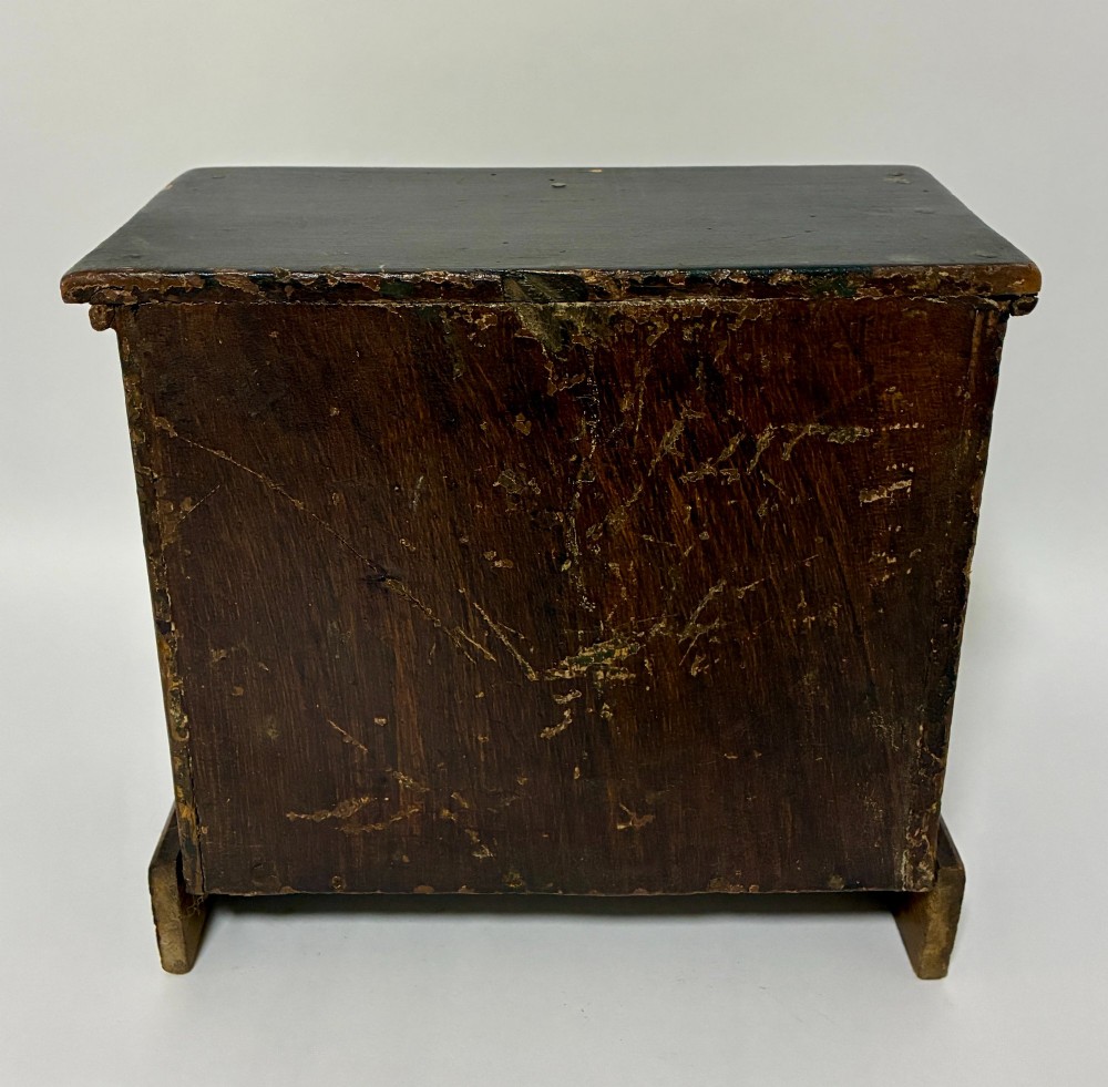 A small wooden miniature chest of drawers featuring two small upper drawers and two full-width lower drawers with turned brass knobs. View V