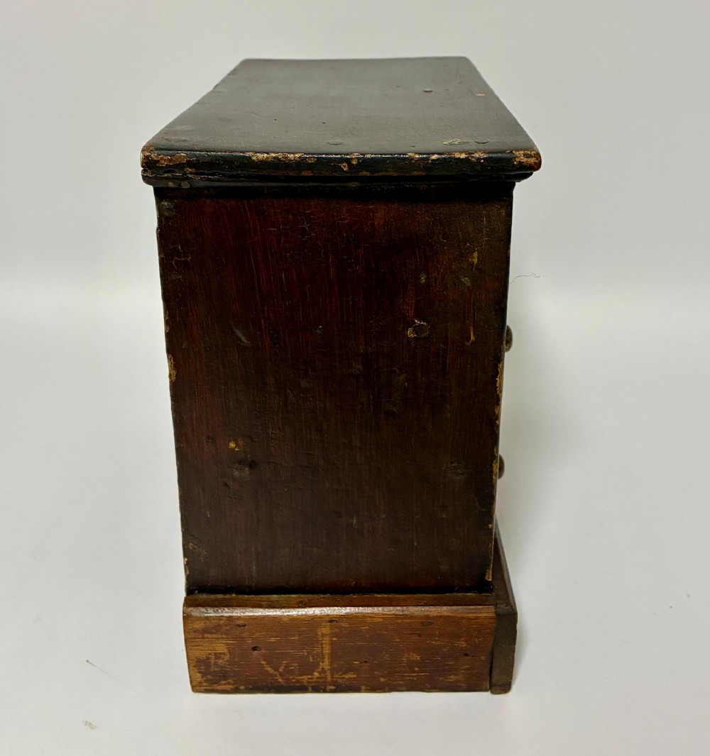 A small wooden miniature chest of drawers featuring two small upper drawers and two full-width lower drawers with turned brass knobs. View VI