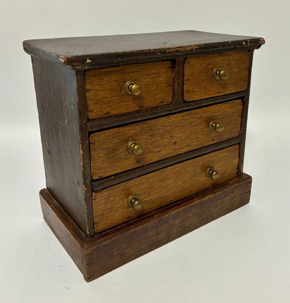A small wooden miniature chest of drawers featuring two small upper drawers and two full-width lower drawers with turned brass knobs. View VII