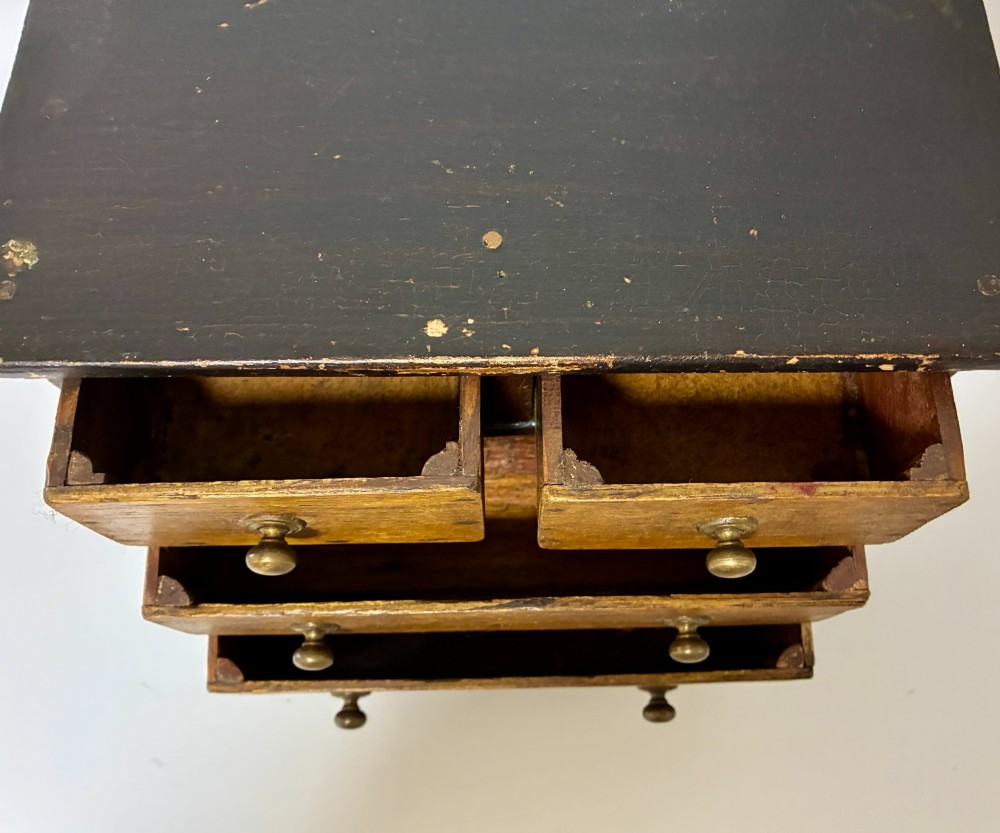 A small wooden miniature chest of drawers featuring two small upper drawers and two full-width lower drawers with turned brass knobs. View IX