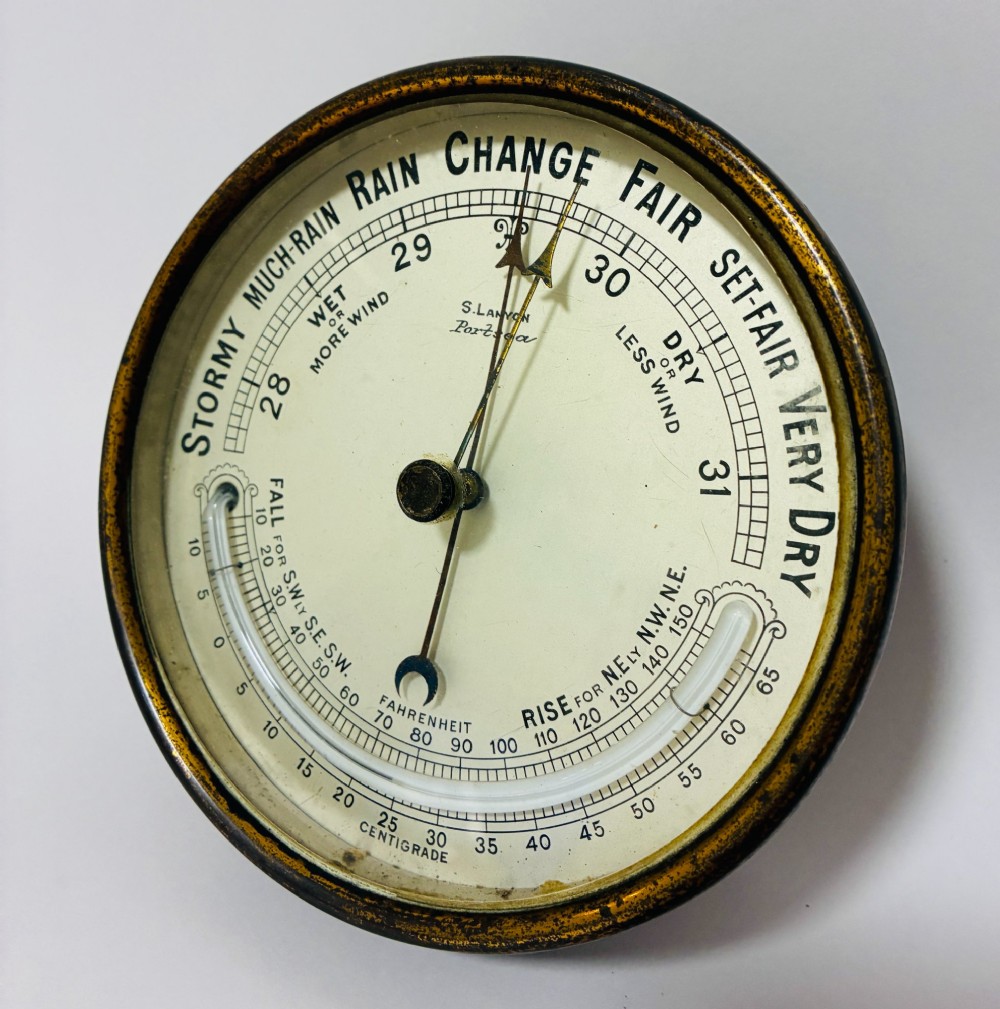 A circular antique brass wall barometer with a cream dial displaying weather conditions and an integrated glass tube thermometer.