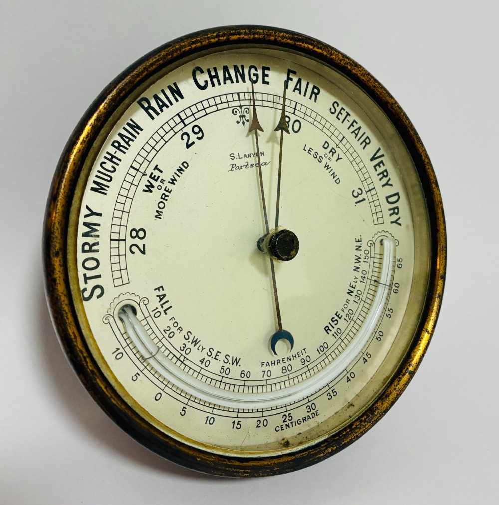 A circular antique brass wall barometer with a cream dial displaying weather conditions and an integrated glass tube thermometer. View II