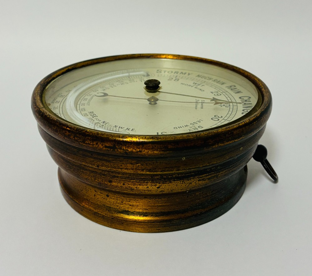 A circular antique brass wall barometer with a cream dial displaying weather conditions and an integrated glass tube thermometer. View V