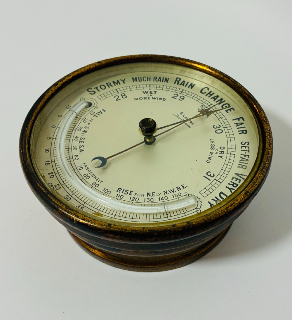 A circular antique brass wall barometer with a cream dial displaying weather conditions and an integrated glass tube thermometer. View VII