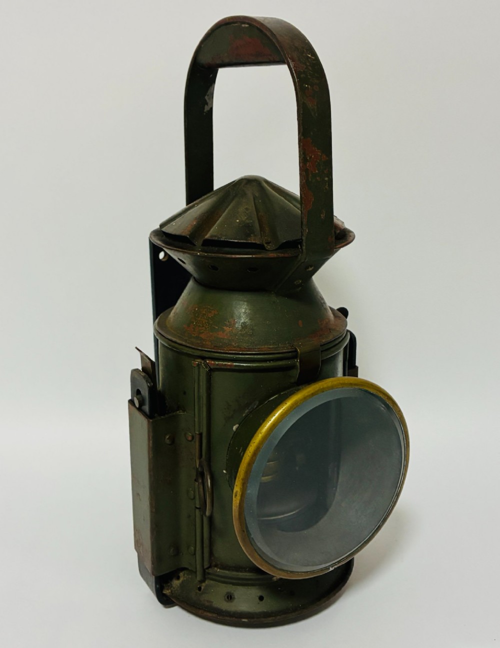 A vintage olive green painted metal railway signal lamp featuring a large round glass lens with a brass rim and a tall arched carrying handle.