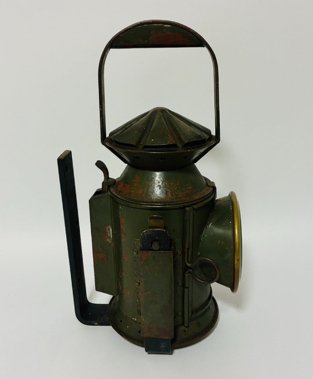 A vintage olive green painted metal railway signal lamp featuring a large round glass lens with a brass rim and a tall arched carrying handle. View II