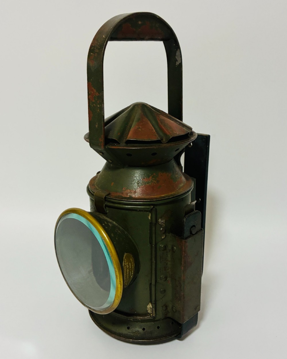 A vintage olive green painted metal railway signal lamp featuring a large round glass lens with a brass rim and a tall arched carrying handle. View VI