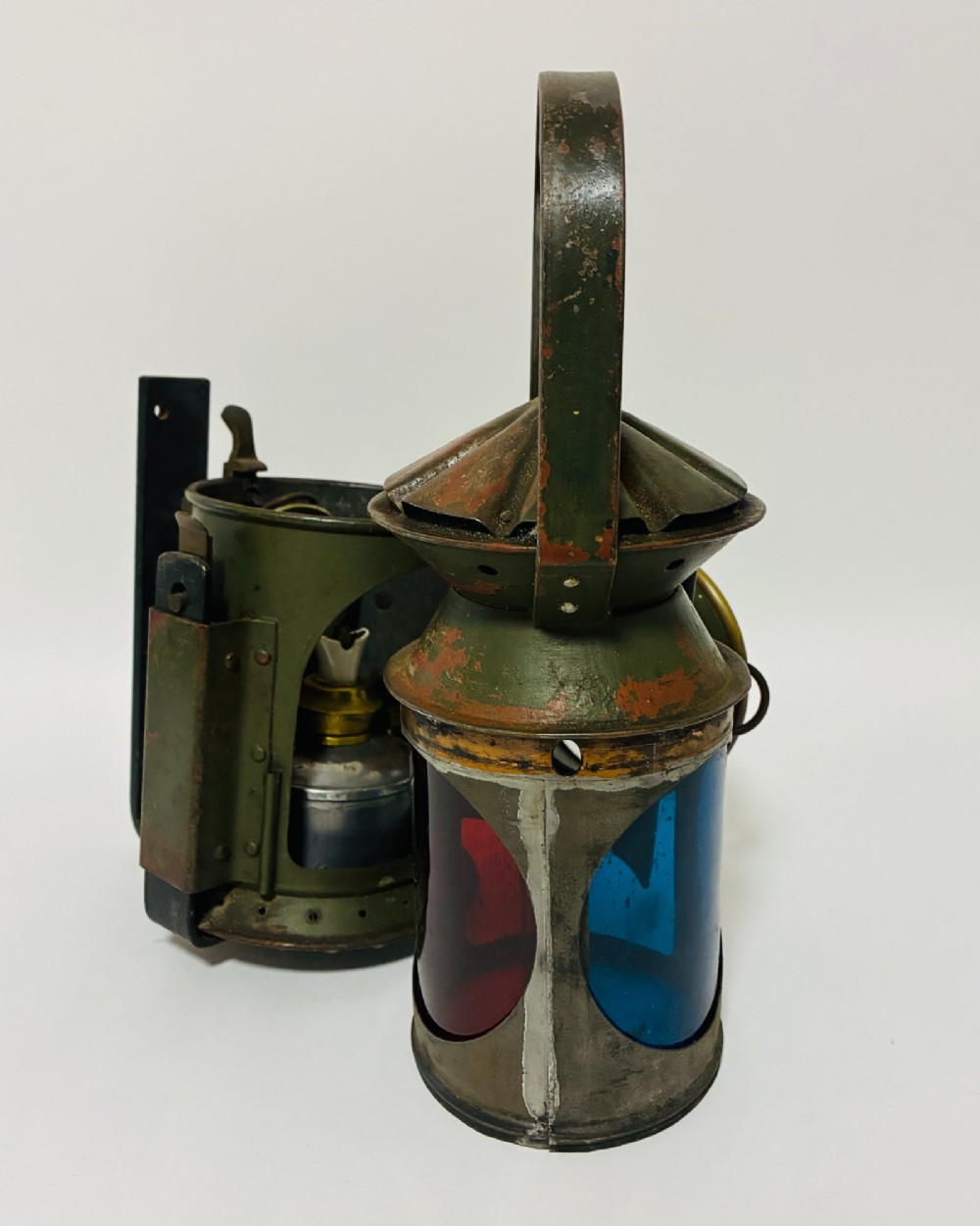 A vintage olive green painted metal railway signal lamp featuring a large round glass lens with a brass rim and a tall arched carrying handle. View X