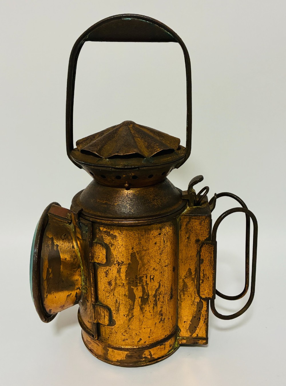 An antique metal railway signalling lamp with an arched top carrying handle, a conical vented cap, and a prominent circular glass lens. View V