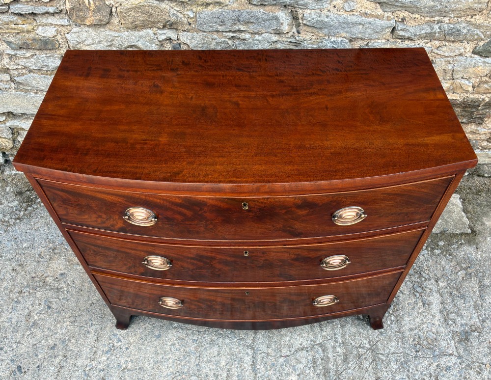 An antique Regency mahogany chest of drawers featuring a curved bowfront shape, three drawers with oval brass handles, and splayed bracket feet. View II
