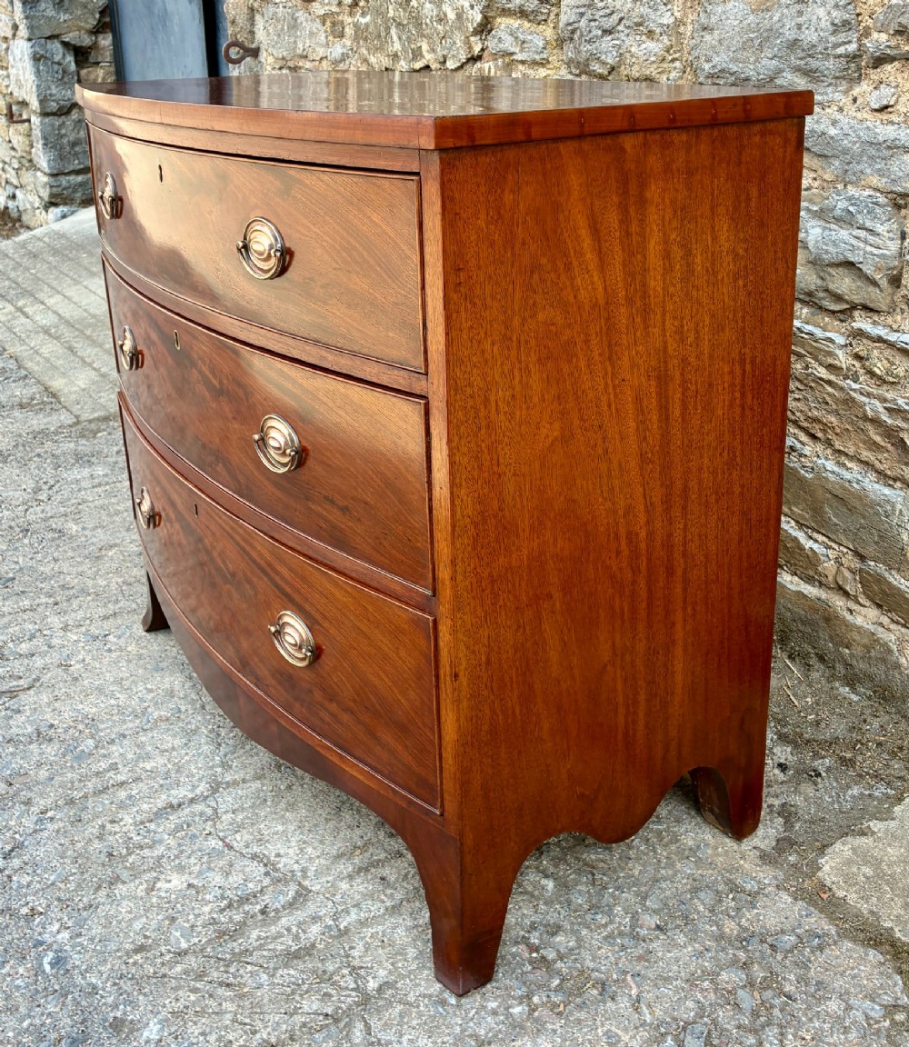 An antique Regency mahogany chest of drawers featuring a curved bowfront shape, three drawers with oval brass handles, and splayed bracket feet. View V