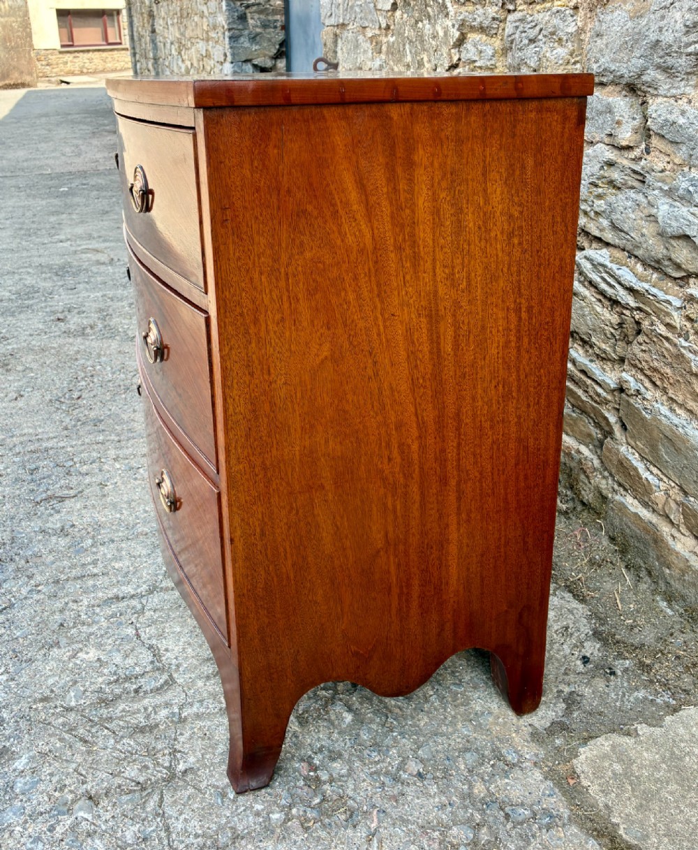 An antique Regency mahogany chest of drawers featuring a curved bowfront shape, three drawers with oval brass handles, and splayed bracket feet. View VI