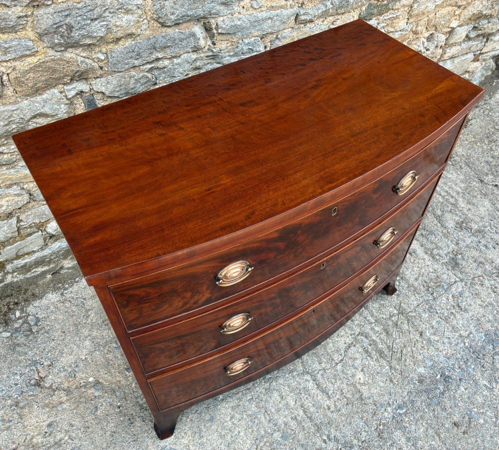 An antique Regency mahogany chest of drawers featuring a curved bowfront shape, three drawers with oval brass handles, and splayed bracket feet. View VII