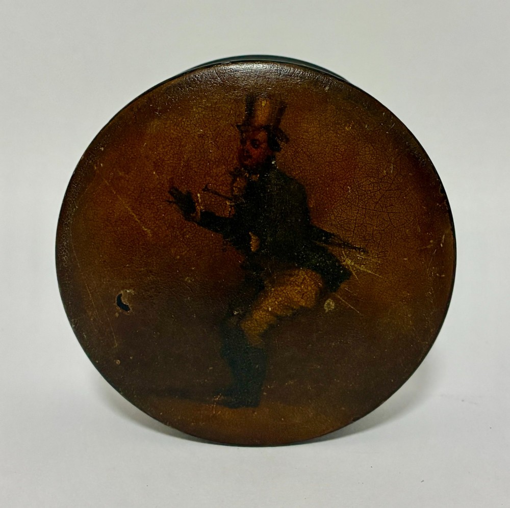 A circular Georgian papier-mache table snuff box featuring a hand-painted depiction of a gentleman in a top hat and tailcoat against a dark lacquered background.