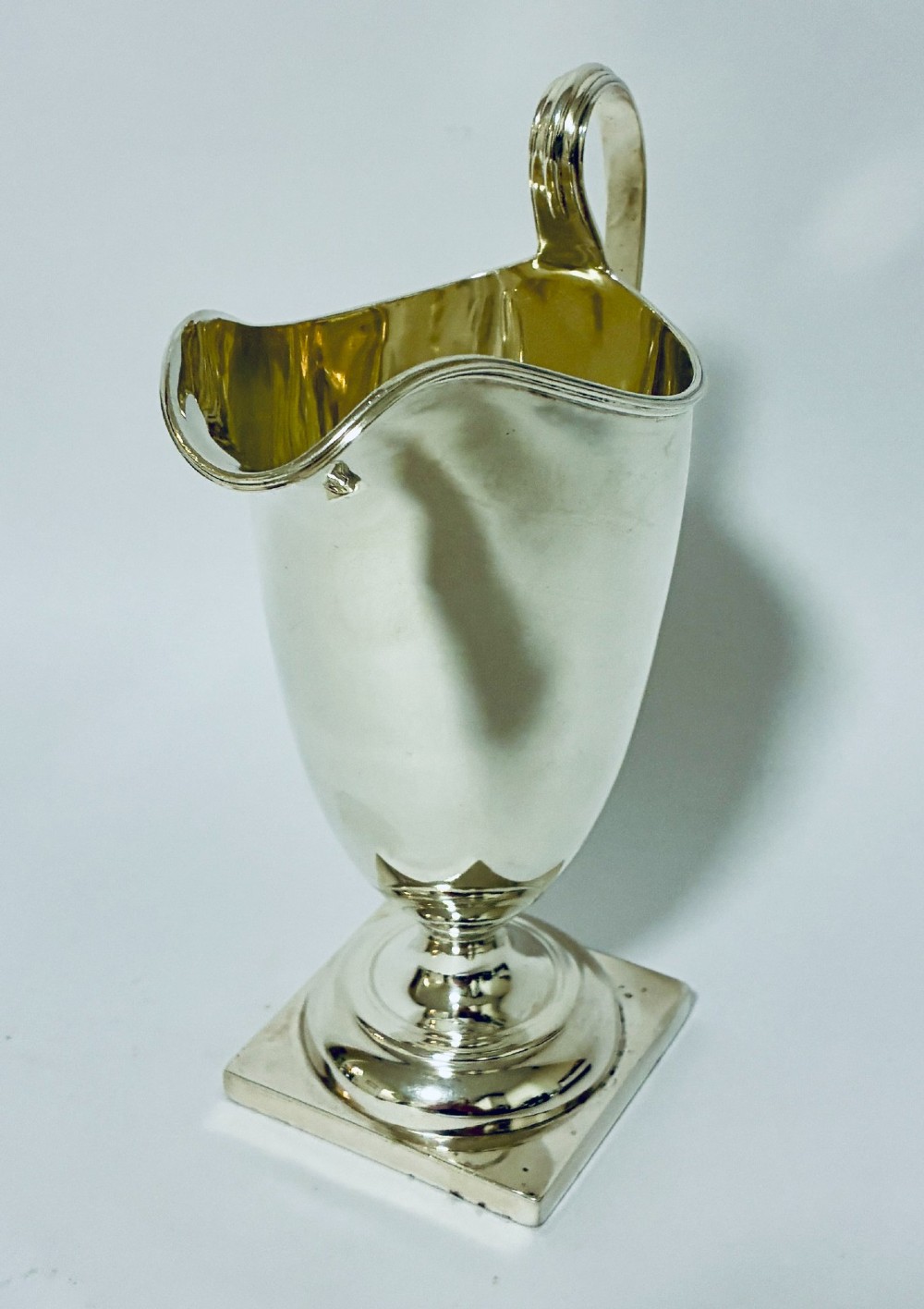 A sterling silver helmet-shaped cream jug featuring a high loop handle, side hallmarks, and a square plinth base. View V