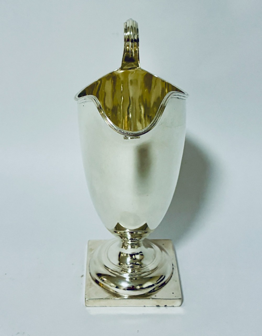 A sterling silver helmet-shaped cream jug featuring a high loop handle, side hallmarks, and a square plinth base. View VI
