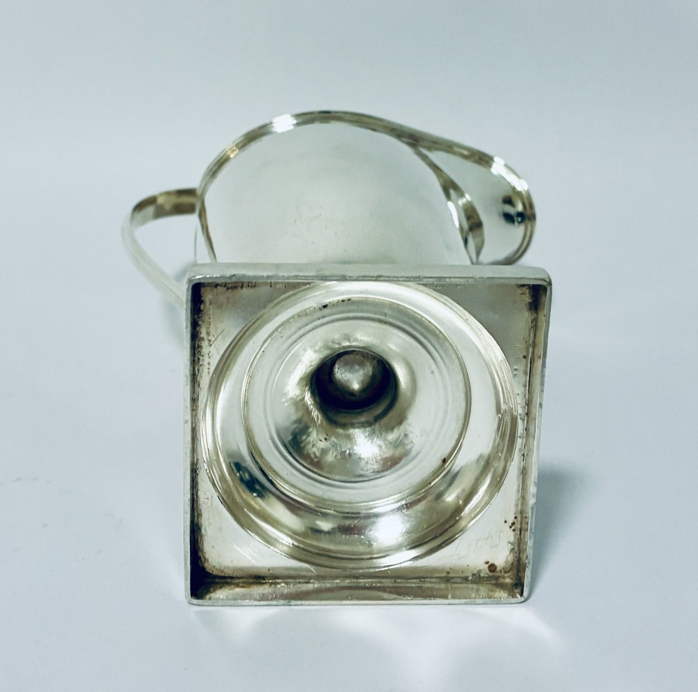A sterling silver helmet-shaped cream jug featuring a high loop handle, side hallmarks, and a square plinth base. View IX