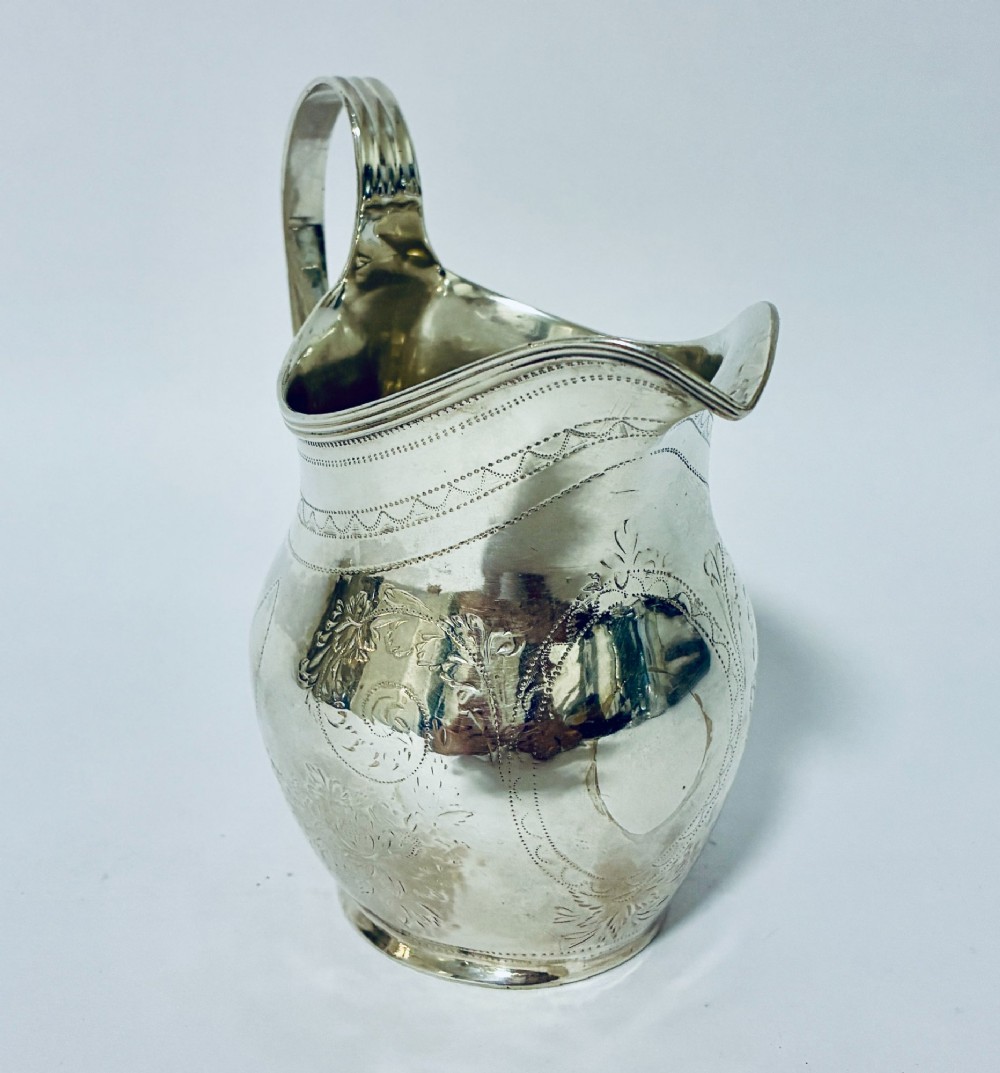 A Georgian sterling silver cream jug featuring a high loop handle, a shaped spout, and fine floral engraving across the body. View II