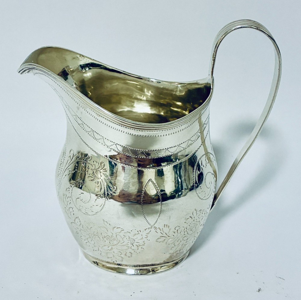 A Georgian sterling silver cream jug featuring a high loop handle, a shaped spout, and fine floral engraving across the body. View IV