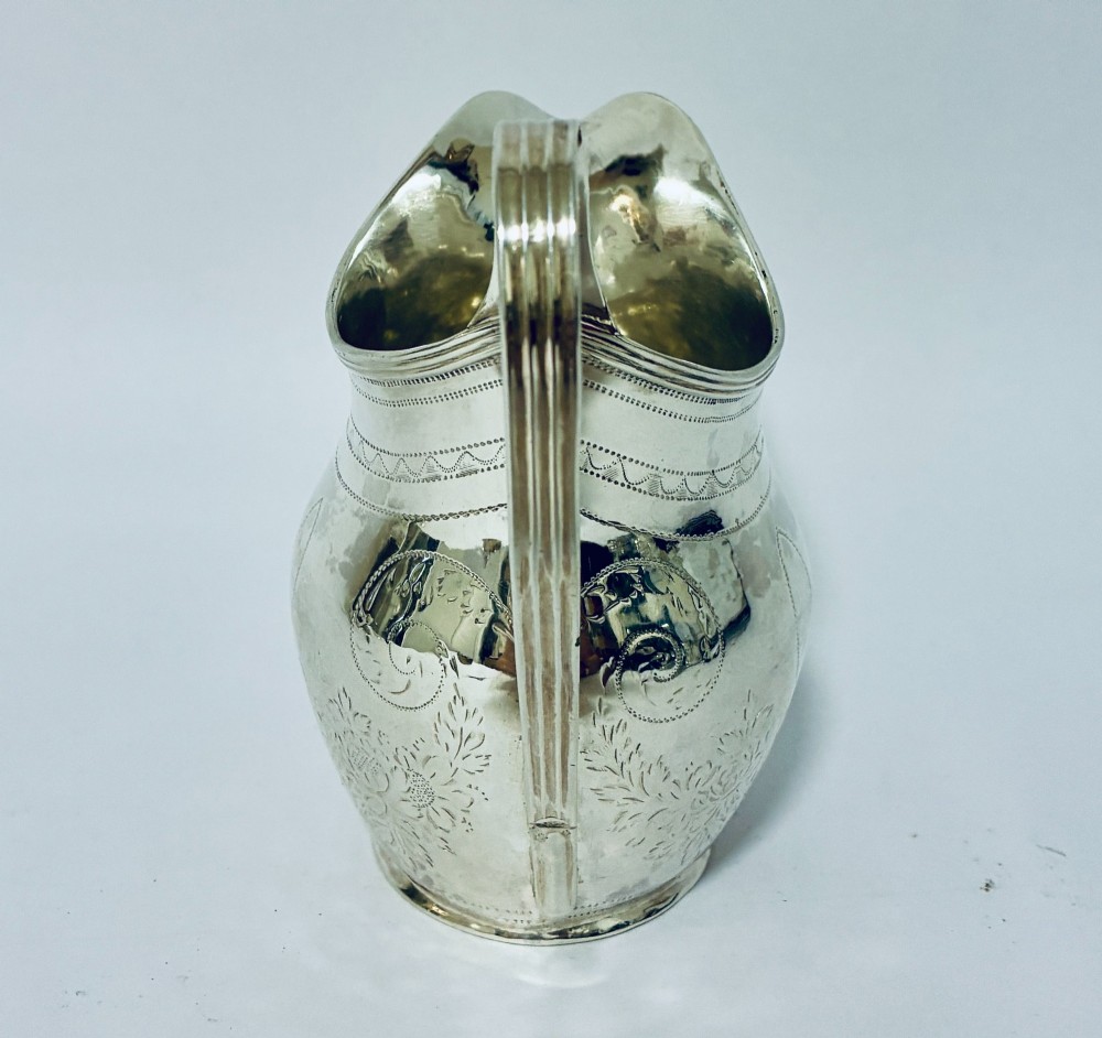 A Georgian sterling silver cream jug featuring a high loop handle, a shaped spout, and fine floral engraving across the body. View V