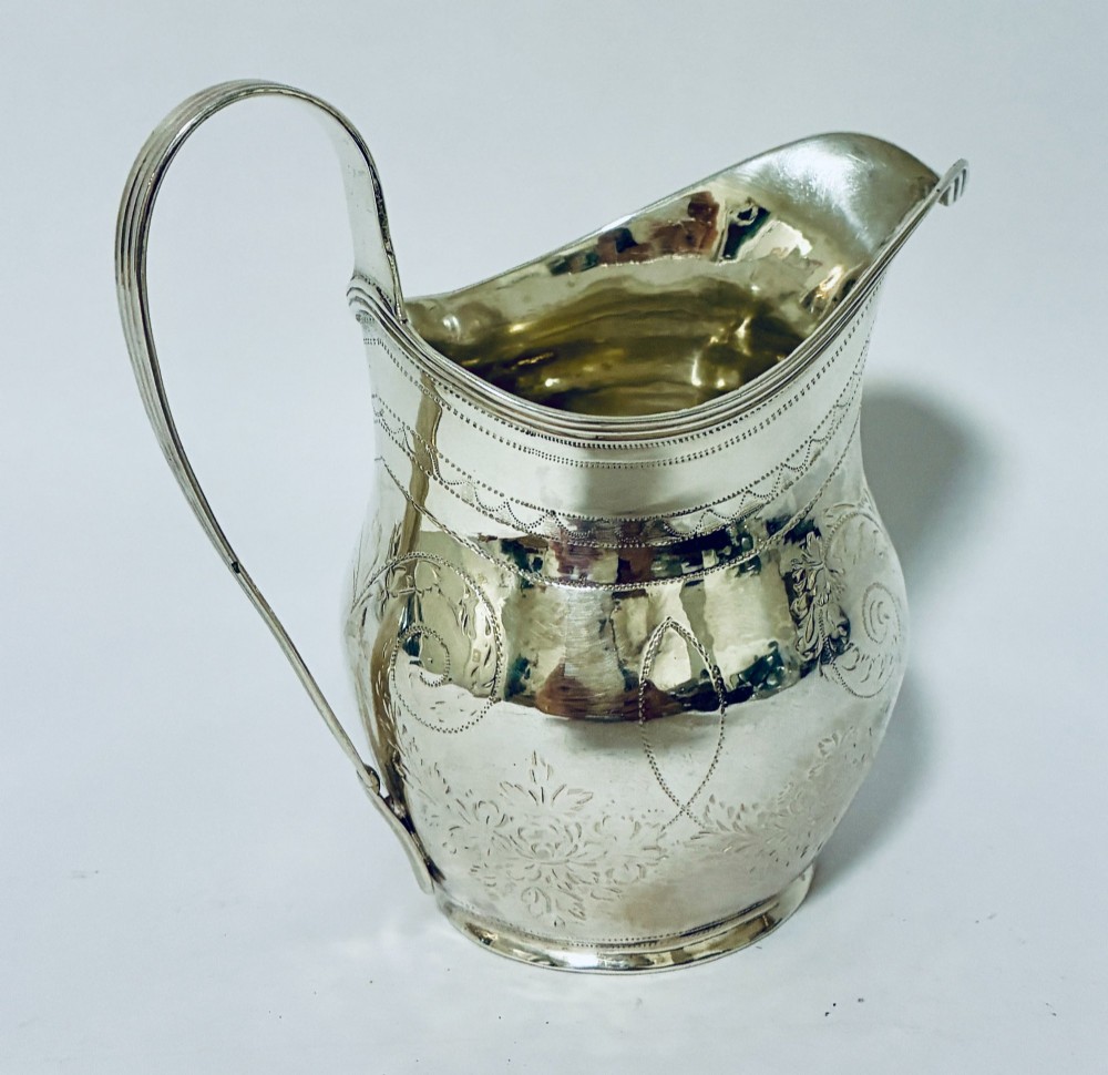 A Georgian sterling silver cream jug featuring a high loop handle, a shaped spout, and fine floral engraving across the body. View VI