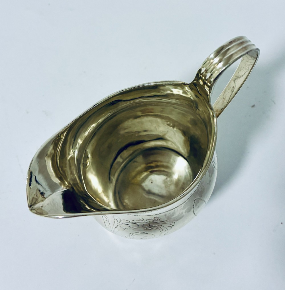 A Georgian sterling silver cream jug featuring a high loop handle, a shaped spout, and fine floral engraving across the body. View IX