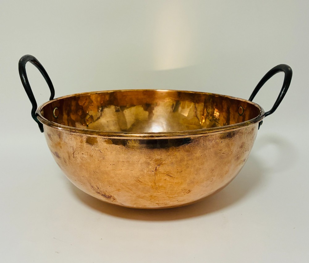 A round antique copper bowl with a wide rim and two black wrought iron handles attached with copper rivets. View II