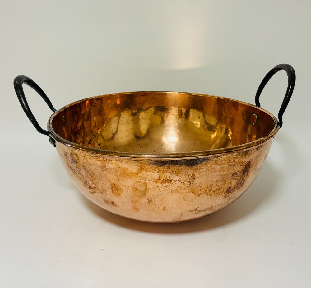 A round antique copper bowl with a wide rim and two black wrought iron handles attached with copper rivets. View V