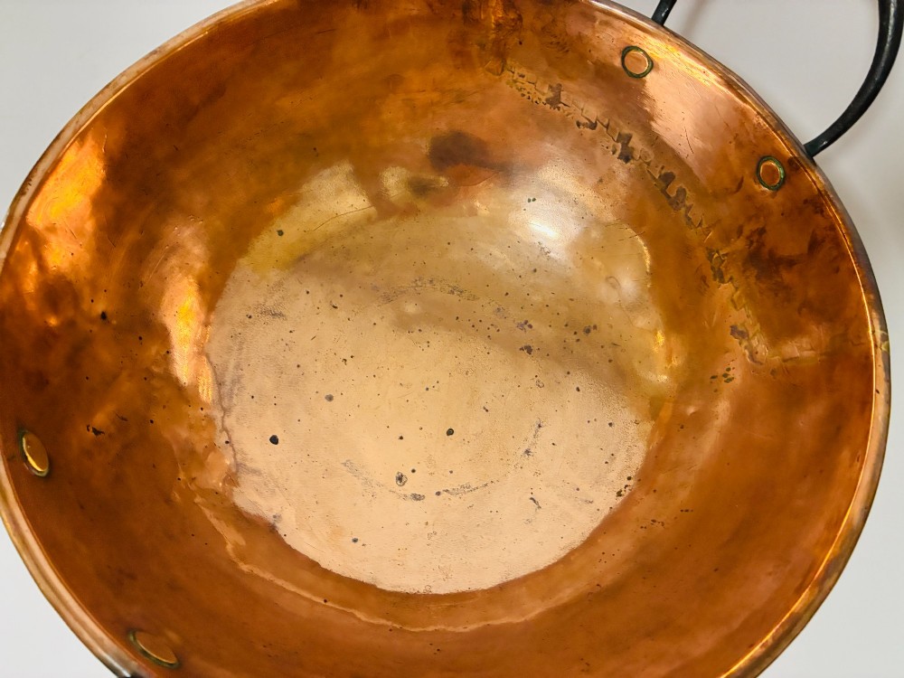 A round antique copper bowl with a wide rim and two black wrought iron handles attached with copper rivets. View X