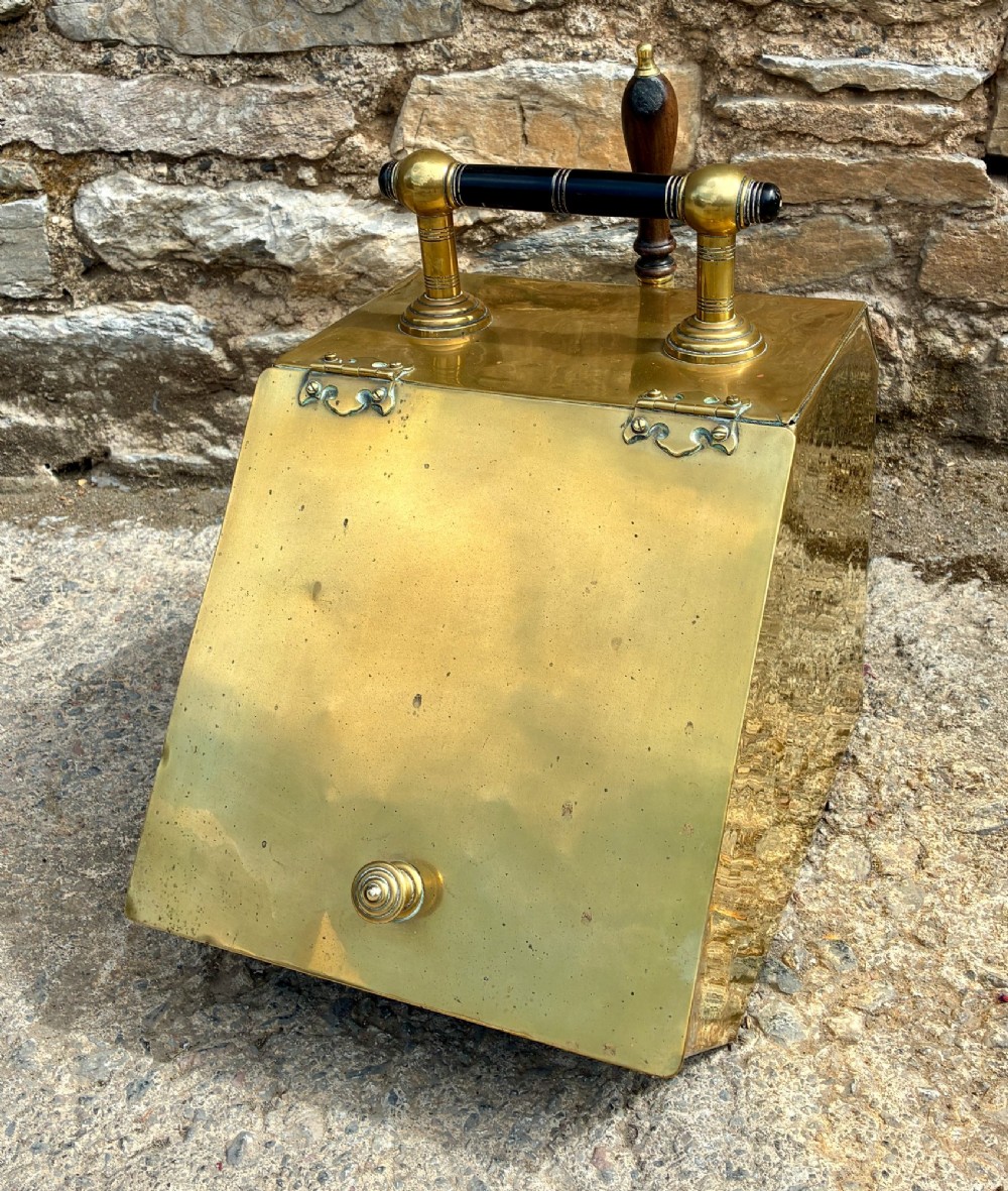 An antique brass sloped coal scuttle featuring a turned wooden carrying handle, a hinged lid, and an attached brass shovel at the rear. View II